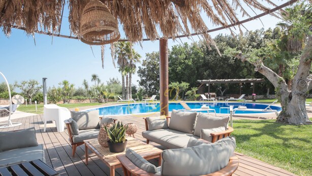 Une terrasse en bois avec des meubles d’extérieur, une piscine et une pergola surmontée d’un toit de chaume.