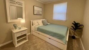 Un dormitorio con cama, mesita de noche, espejo, un cuadro en la pared, ventana con persianas y una planta.
