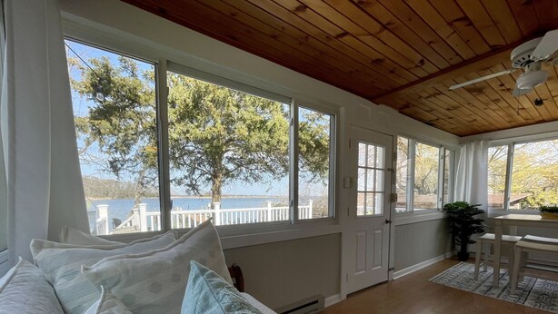 Una habitación acogedora con techo de madera, amplias ventanas con vistas a un lago, un sofá blanco con cojines estampados y una zona de comedor con mesa y sillas.