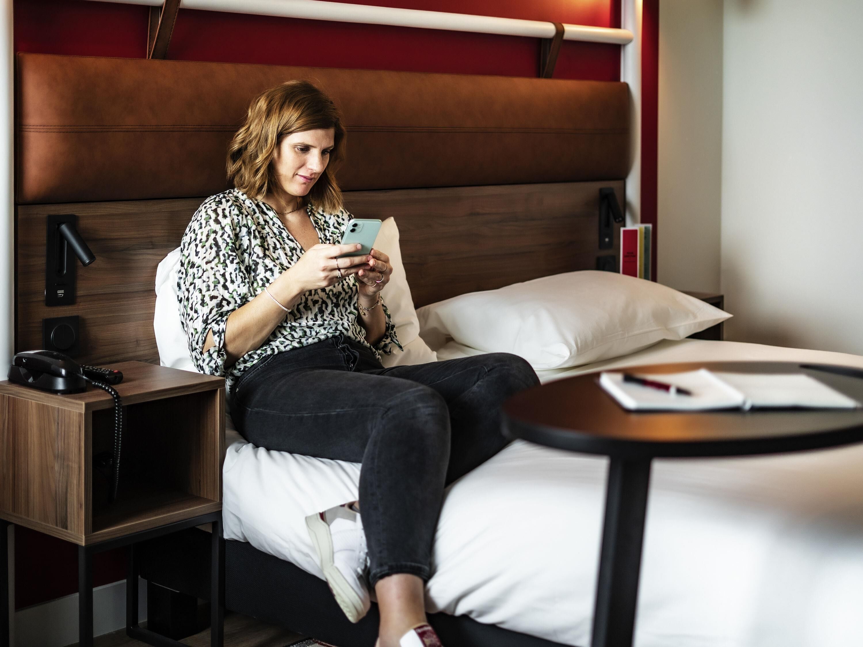 A person sitting on a bed in a hotel room, using a smartphone.