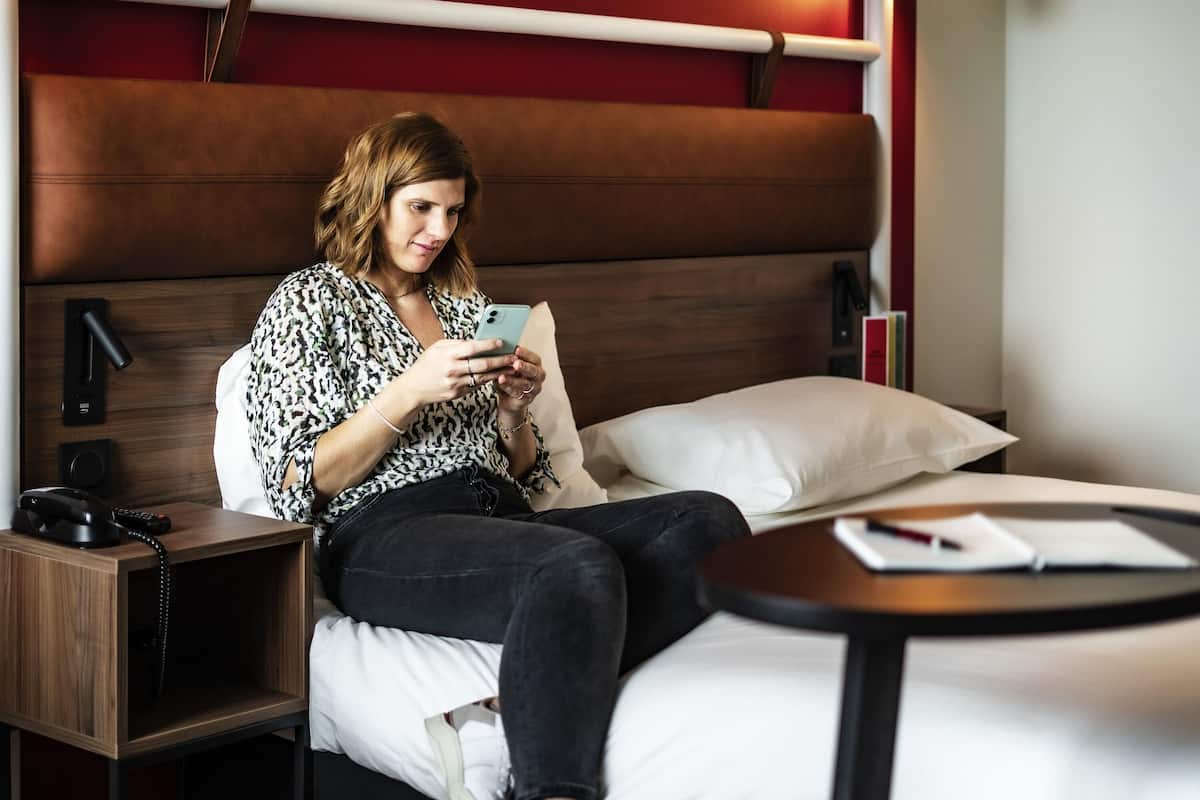 A person sitting on a bed in a hotel room, using a smartphone.