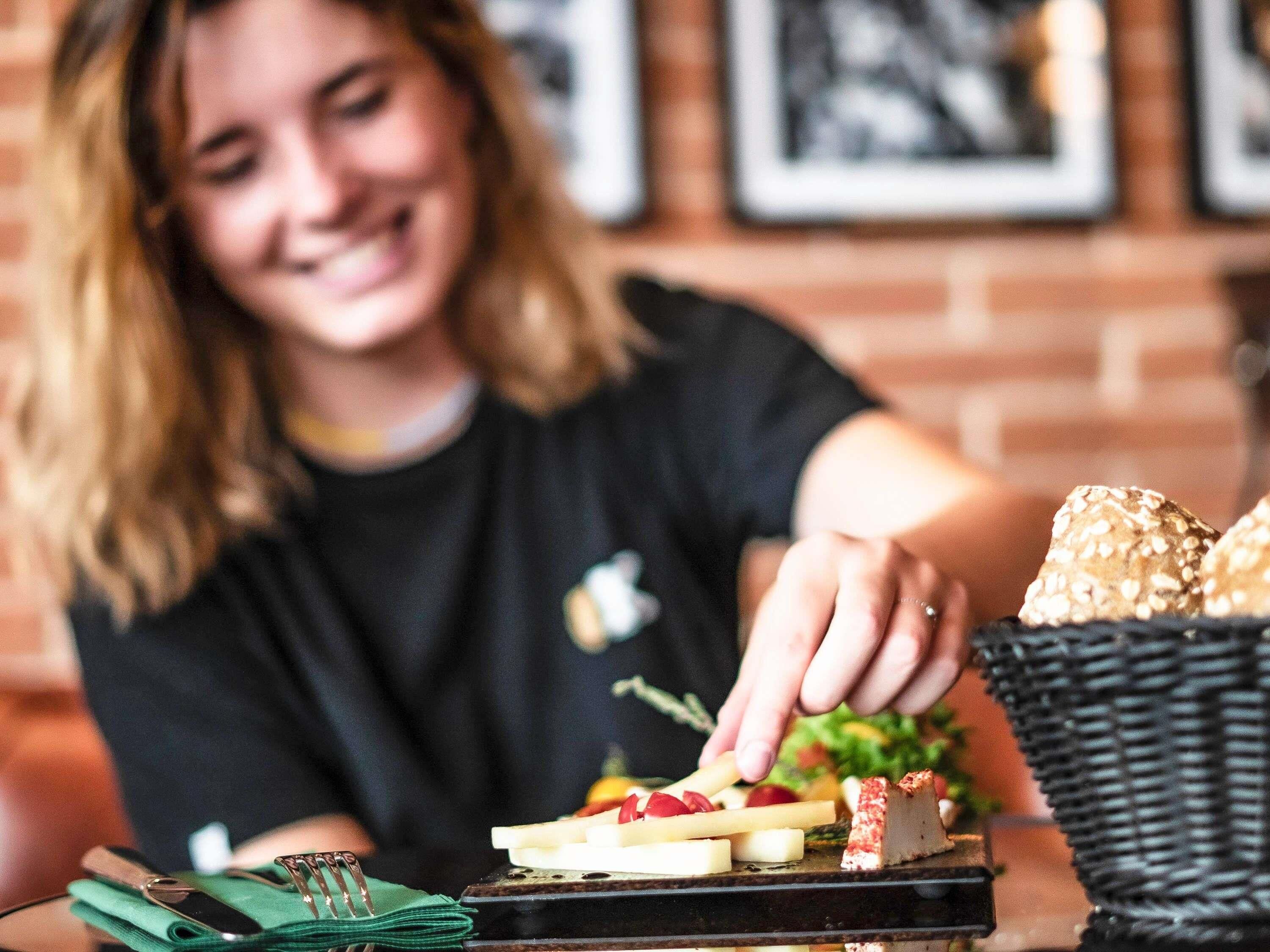 Una persona sentada a una mesa con diversos alimentos, como queso y pan, en un ambiente con pared de ladrillo y cuadros.