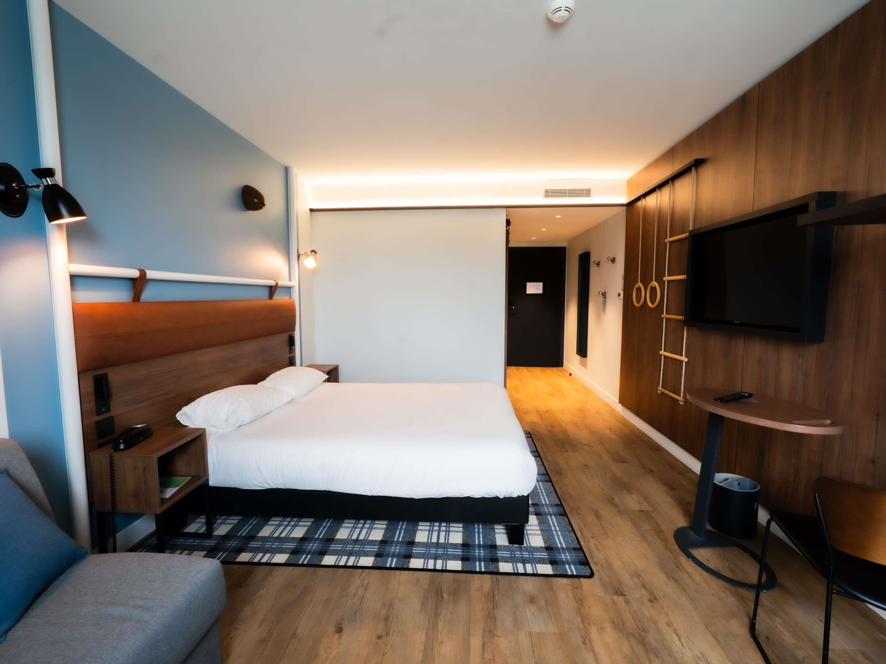 A modern hotel room with a large bed, bedside tables, a TV, and a wooden wardrobe.