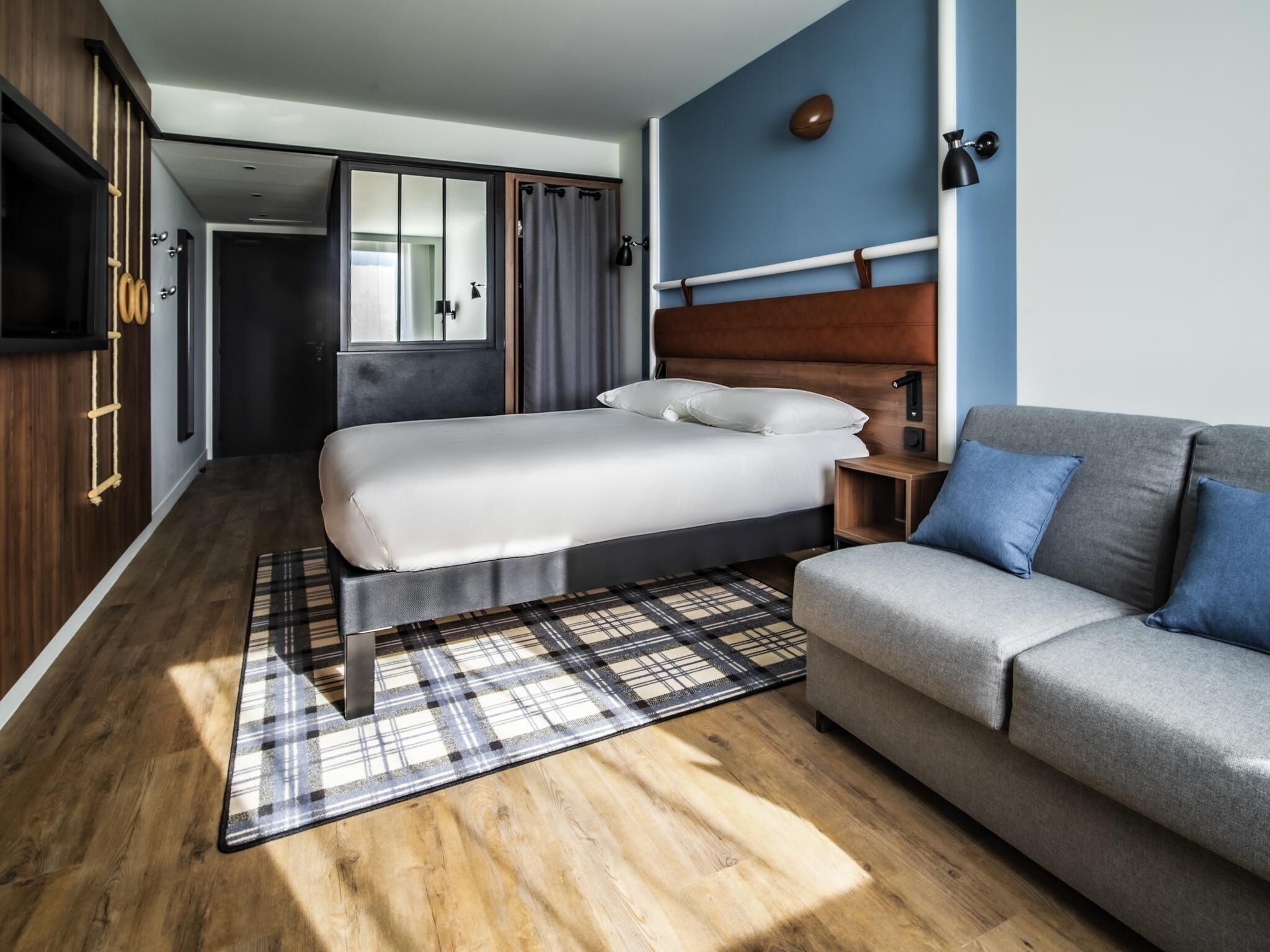 A bedroom with a bed, a sofa, a wooden floor, a plaid rug, and a wardrobe.