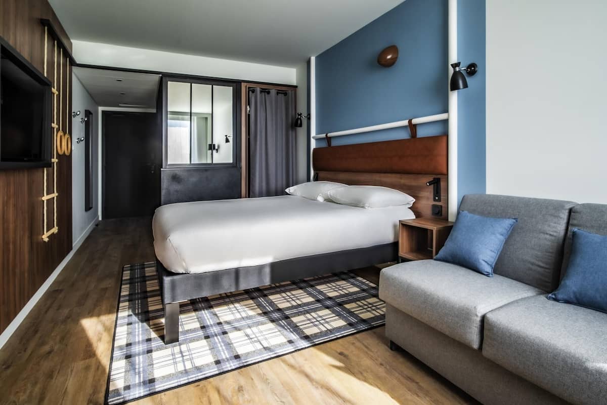 A bedroom with a bed, a sofa, a wooden floor, a plaid rug, and a wardrobe.