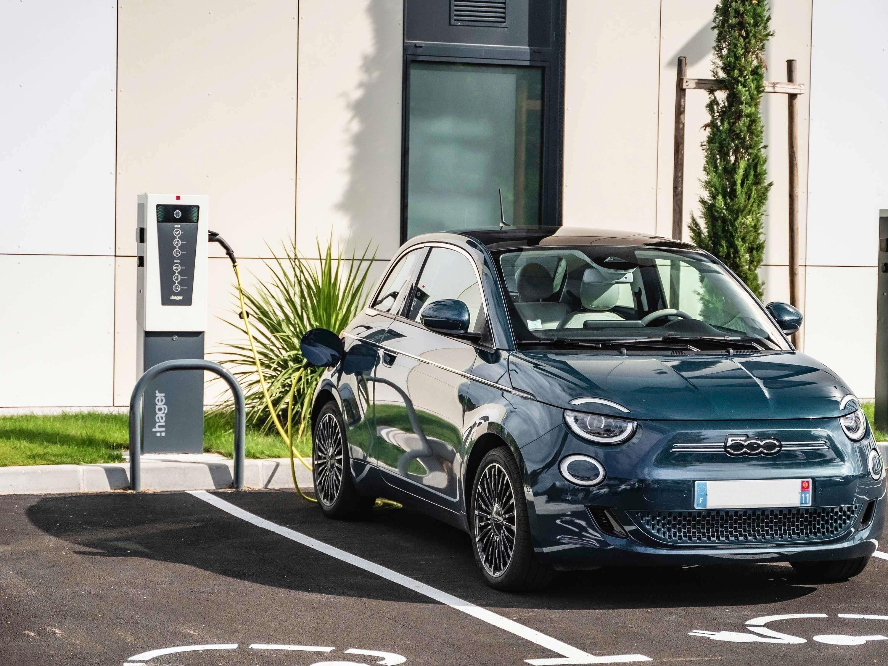 Een kleine auto geparkeerd bij een laadstation voor elektrische auto's, met een gebouw met een modern ontwerp en een heldere lucht.