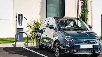 Een kleine auto geparkeerd bij een laadstation voor elektrische auto's, met een gebouw met een modern ontwerp en een heldere lucht.