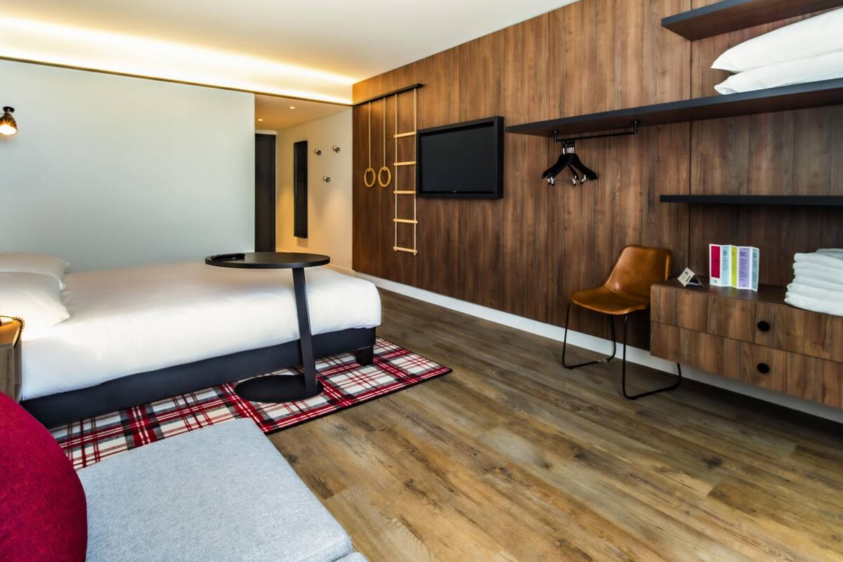 A modern hotel room with a bed, a small round table, a TV mounted on the wall, a wooden wardrobe, and a red plaid rug.