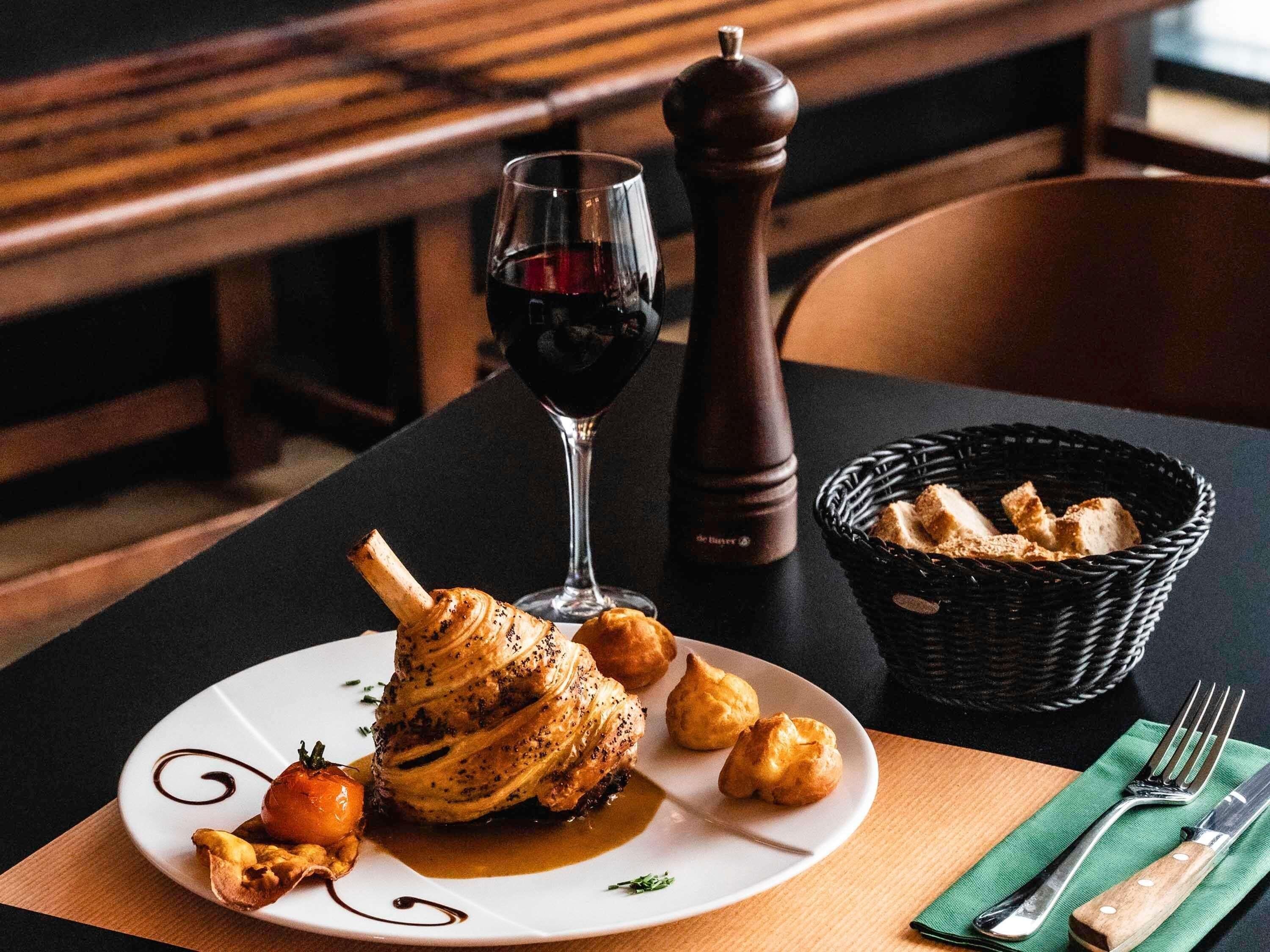 A dining table with a plate of food, a glass of red wine, a pepper mill, and a basket of bread.