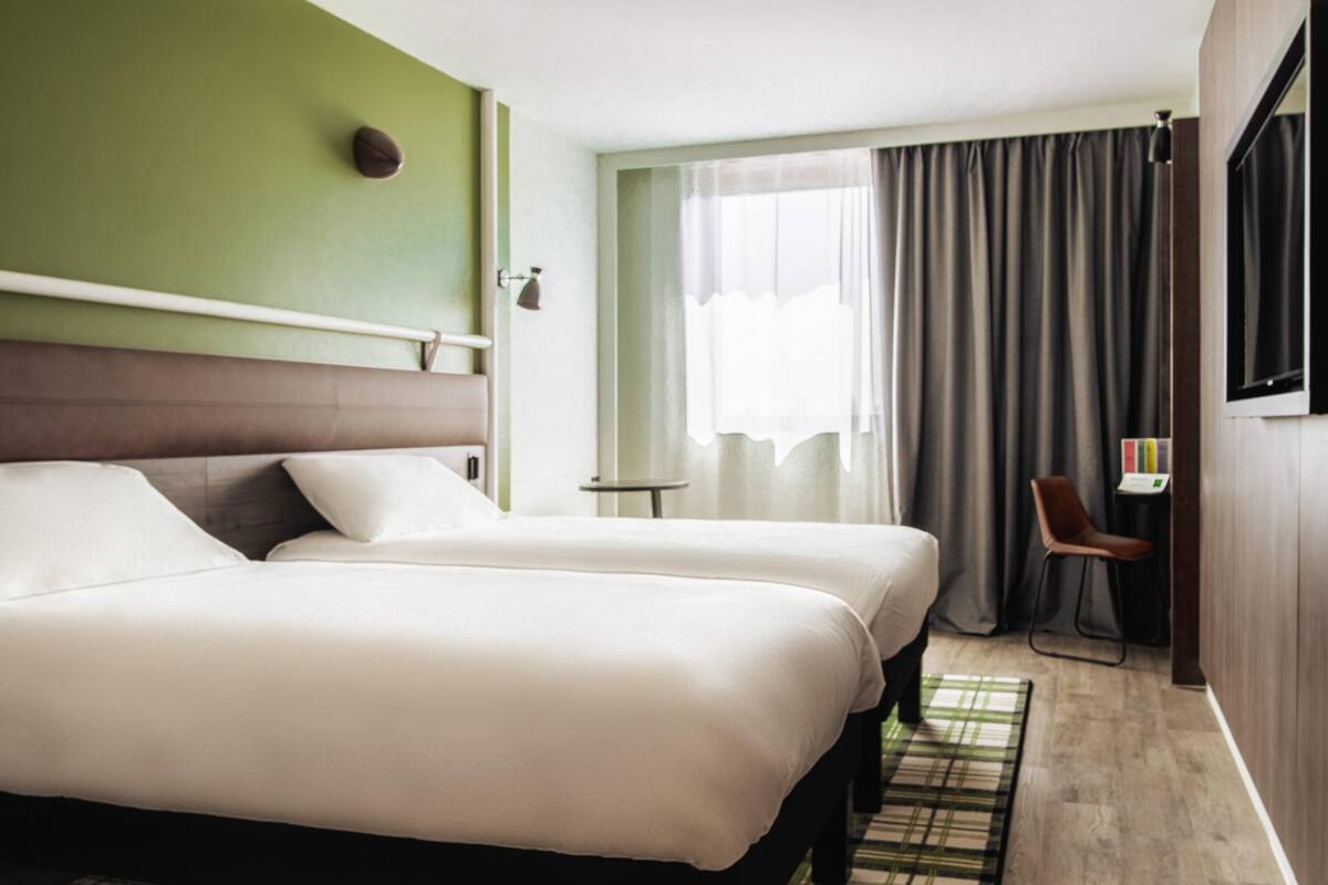 A hotel room with two beds, a small table, a chair, a TV, and a window with curtains.