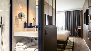 A modern hotel room with a shower, a large mirror, a bed, a bedside table, a TV, and a view of the outdoors. - ibis Styles Castelnaudary (Castelnaudary)