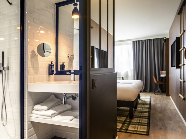 A modern hotel room with a shower, a large mirror, a bed, a bedside table, a TV, and a view of the outdoors. - ibis Styles Castelnaudary (Castelnaudary)