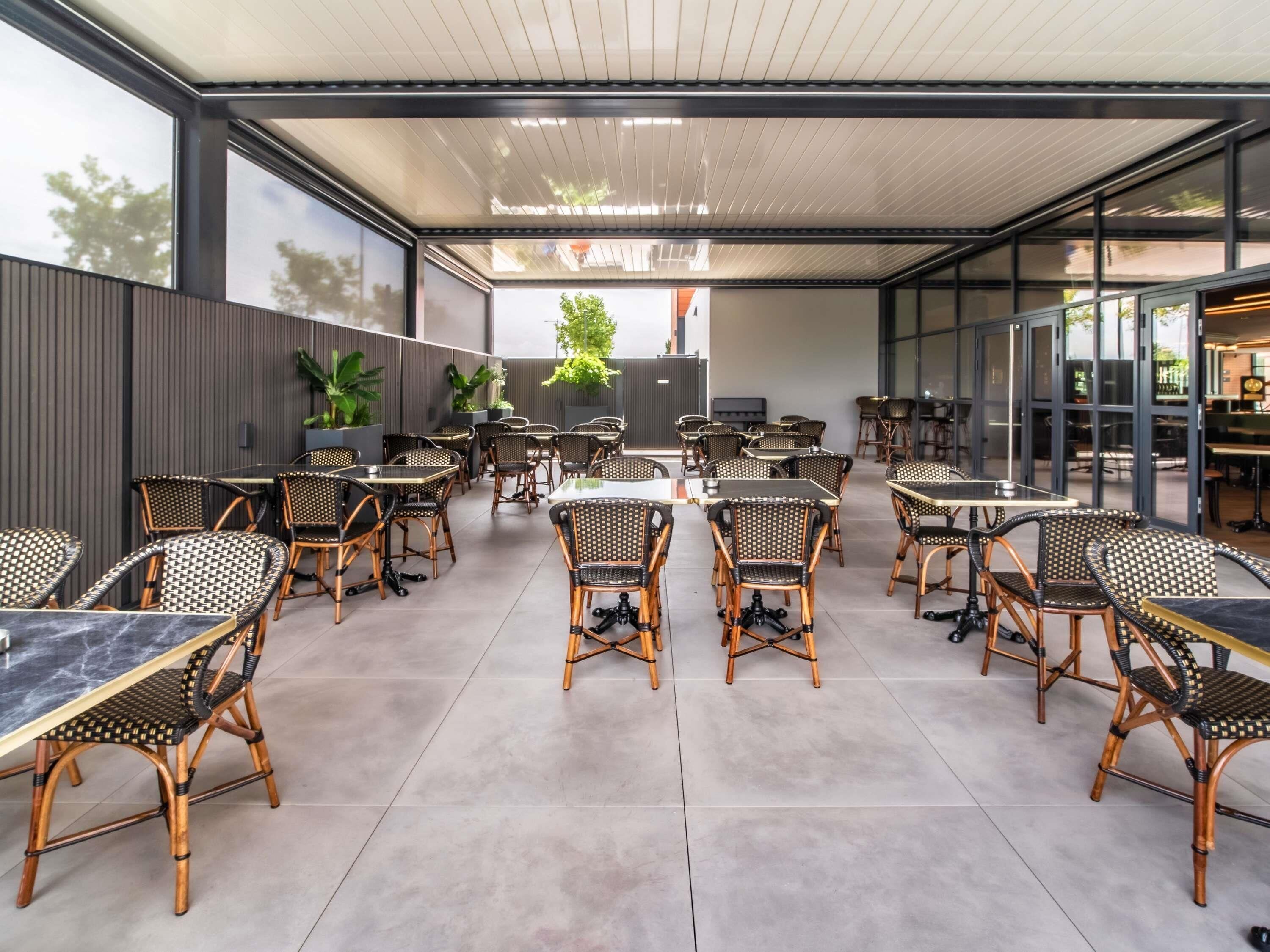 An outdoor dining area with wicker chairs and tables, large windows, and potted plants.