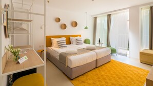 Habitación de hotel moderna con una cama doble, un escritorio y una silla. Se ve una ventana con cortinas, una pequeña mesa con una botella y una lámpara verde.