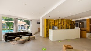 Un moderno vestíbulo de hotel con un mostrador de recepción, una zona de piscina visible a través de grandes ventanales y un área de descanso con sofás y otomanas.