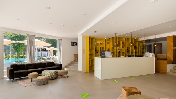 En modern hotellobby med en receptionsdisk, ett poolområde som syns genom stora fönster och en bekväm sittgrupp med en soffa och sittpallar.