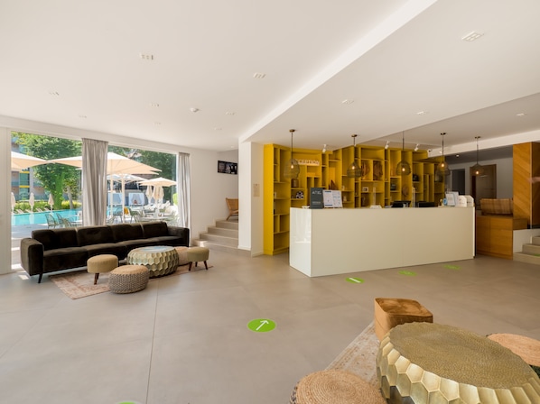 Eine moderne Hotelhalle mit einer Rezeption, einem durch große Fenster sichtbaren Poolbereich und einer gemütlichen Sitzecke mit einer Couch und Hockern.