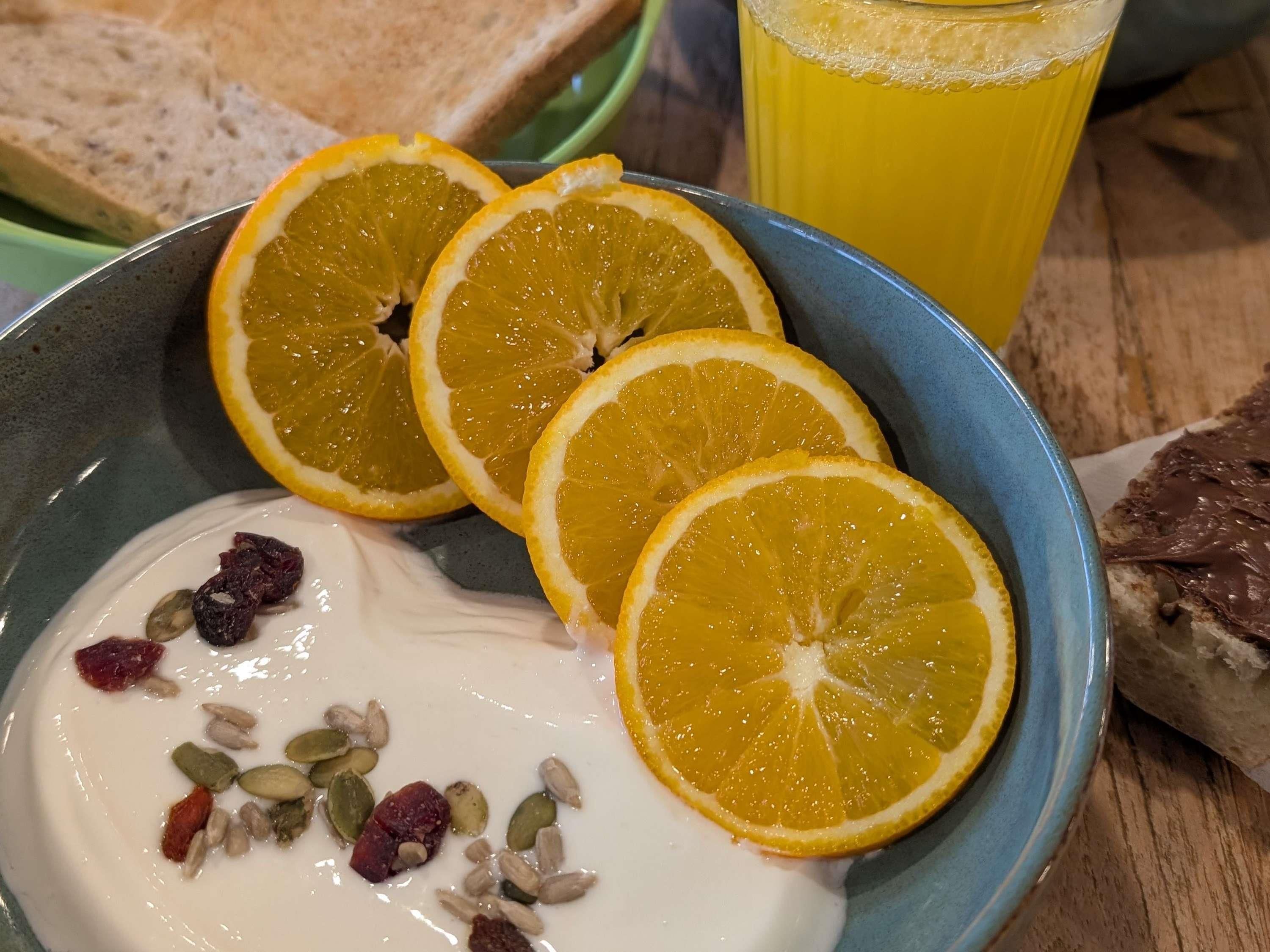A bowl of yogurt with dried fruits and orange slices, a glass of orange juice, and a slice of bread on a wooden table.