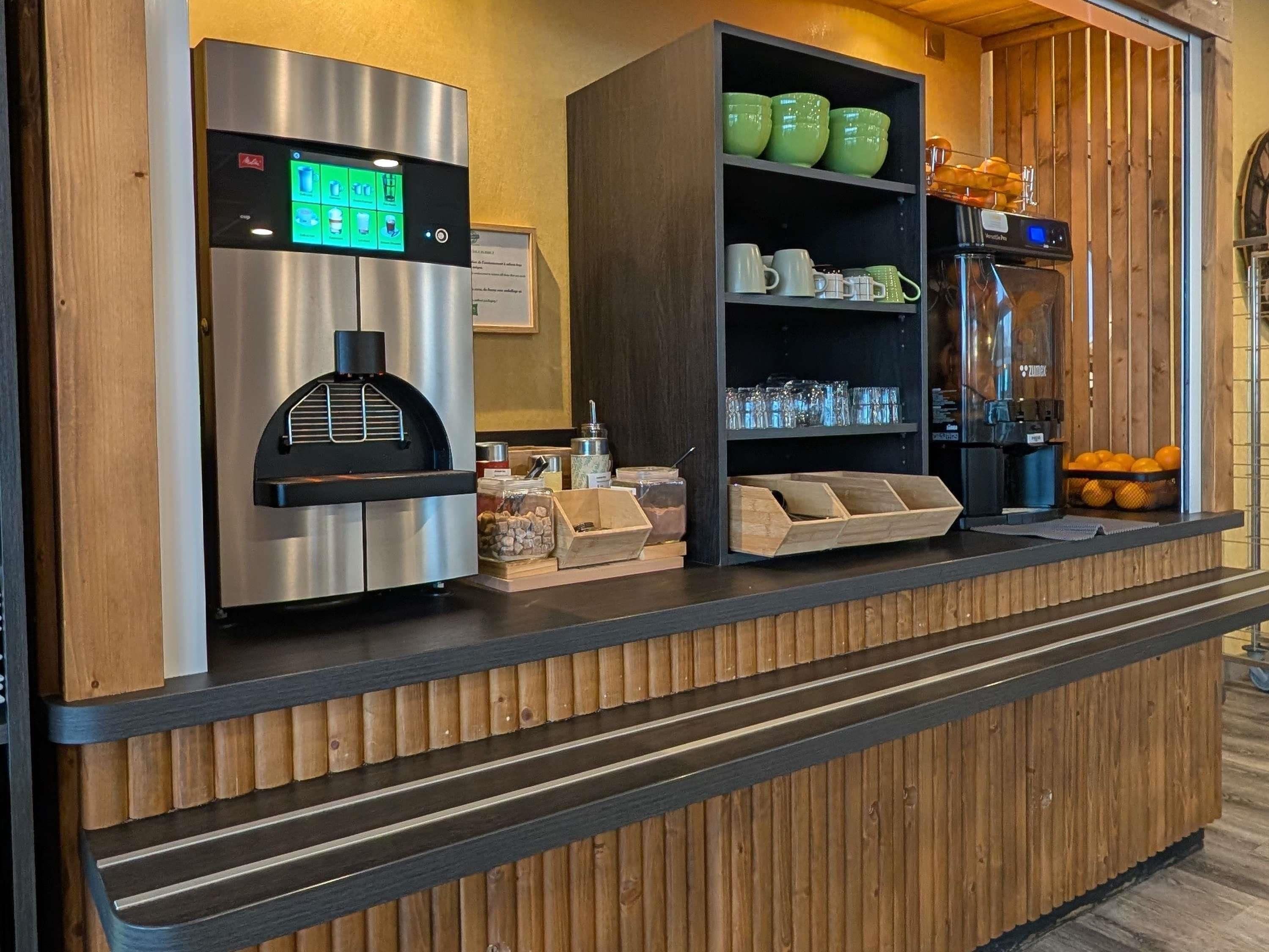 A self-service coffee station with a digital coffee machine, a coffee grinder, and a variety of mugs and cups.