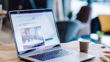 A laptop on a wooden table with a cup of coffee, in a room with large windows and modern furniture.