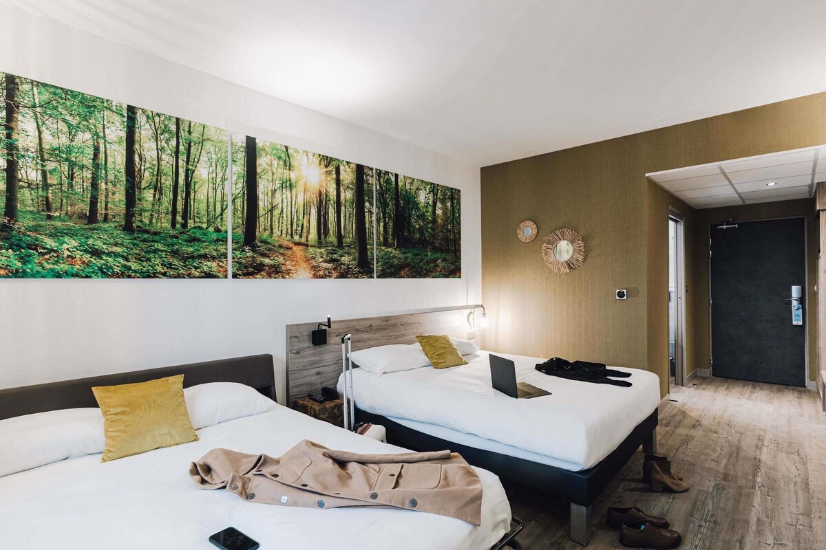Une chambre d’hôtel avec deux lits, une grande fresque murale sur le thème de la forêt, un miroir et une porte donnant sur le couloir.