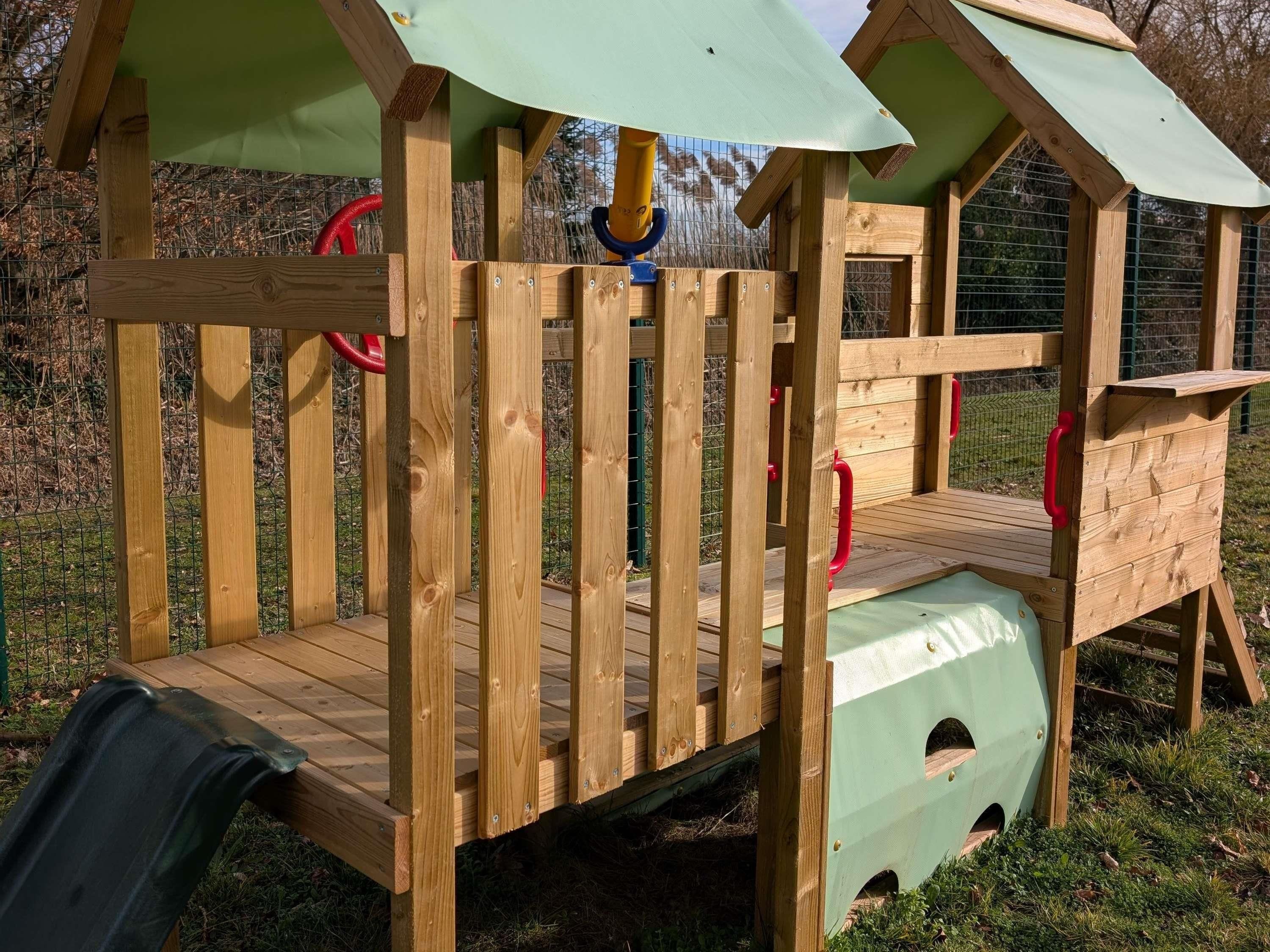 Una casita de juegos de madera con tobogán, zona de escalada y un espacio cubierto.