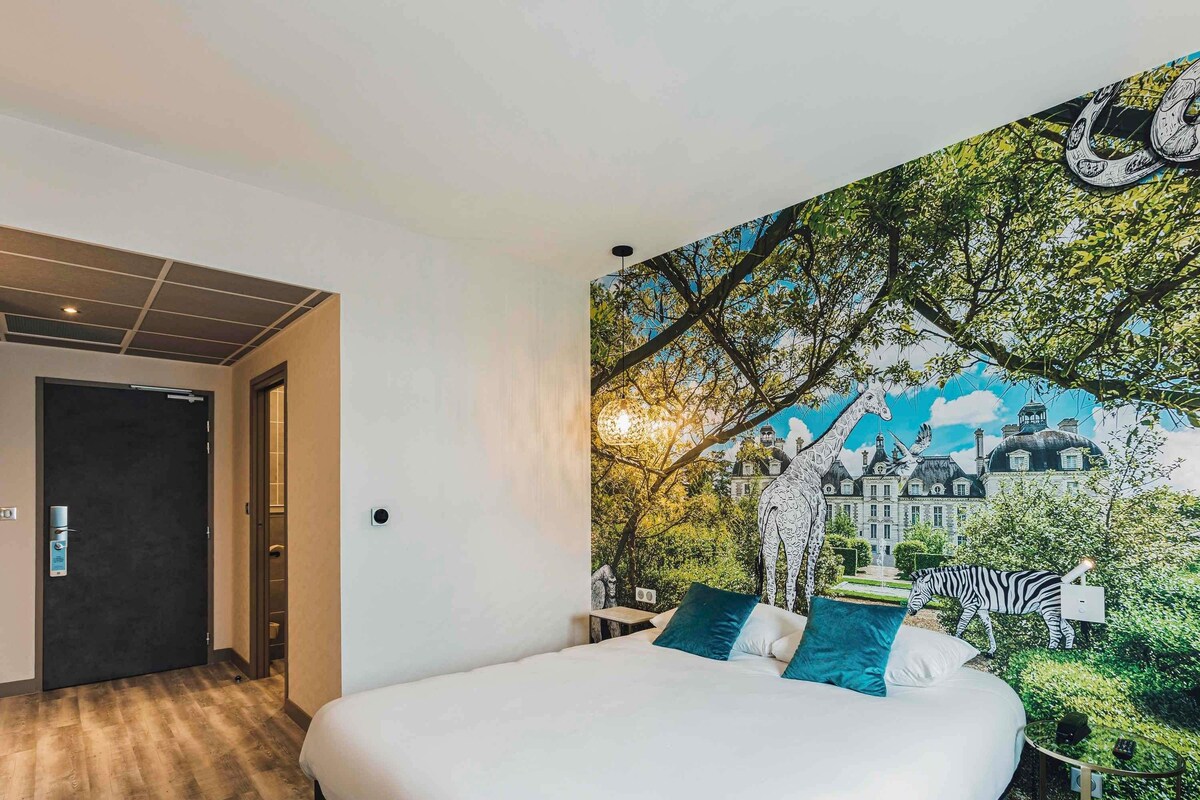 Une chambre avec un grand lit, une porte et une peinture murale représentant un bâtiment et une girafe.