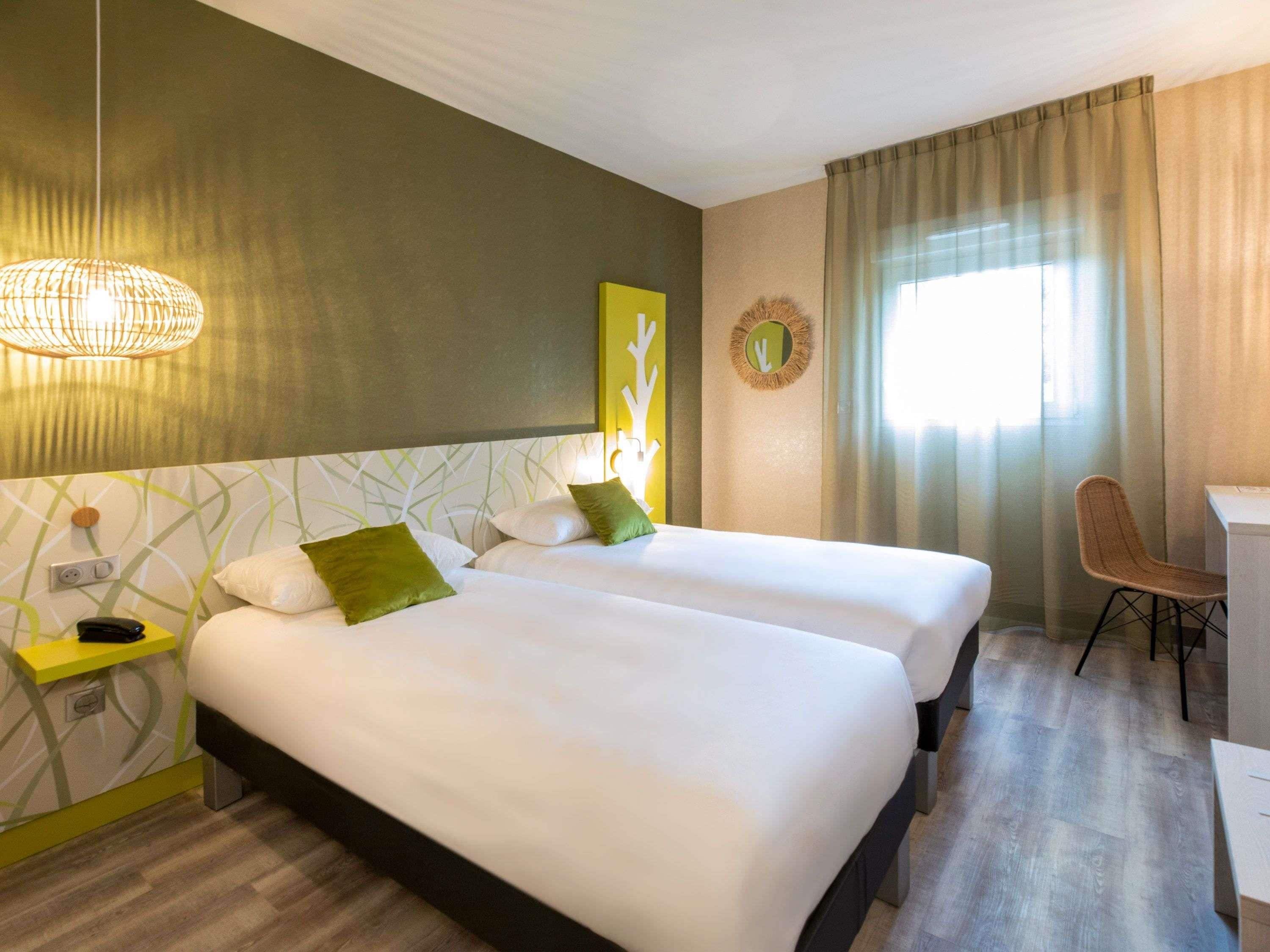 A hotel room with two beds, a desk, a chair, a lamp, and a window with curtains.