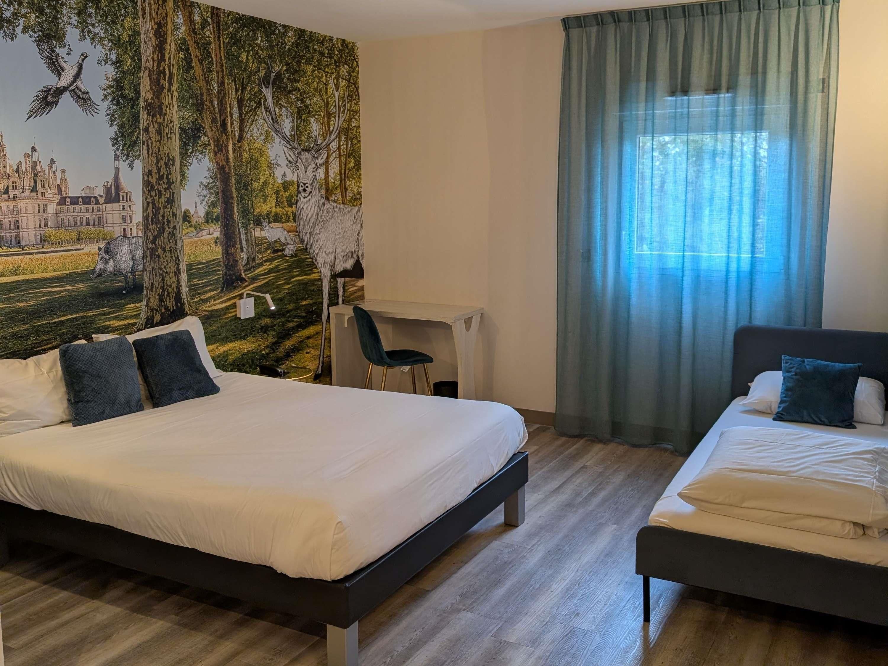 Habitación de hotel con dos camas, un escritorio, una silla y un gran mural con un paisaje de bosque.