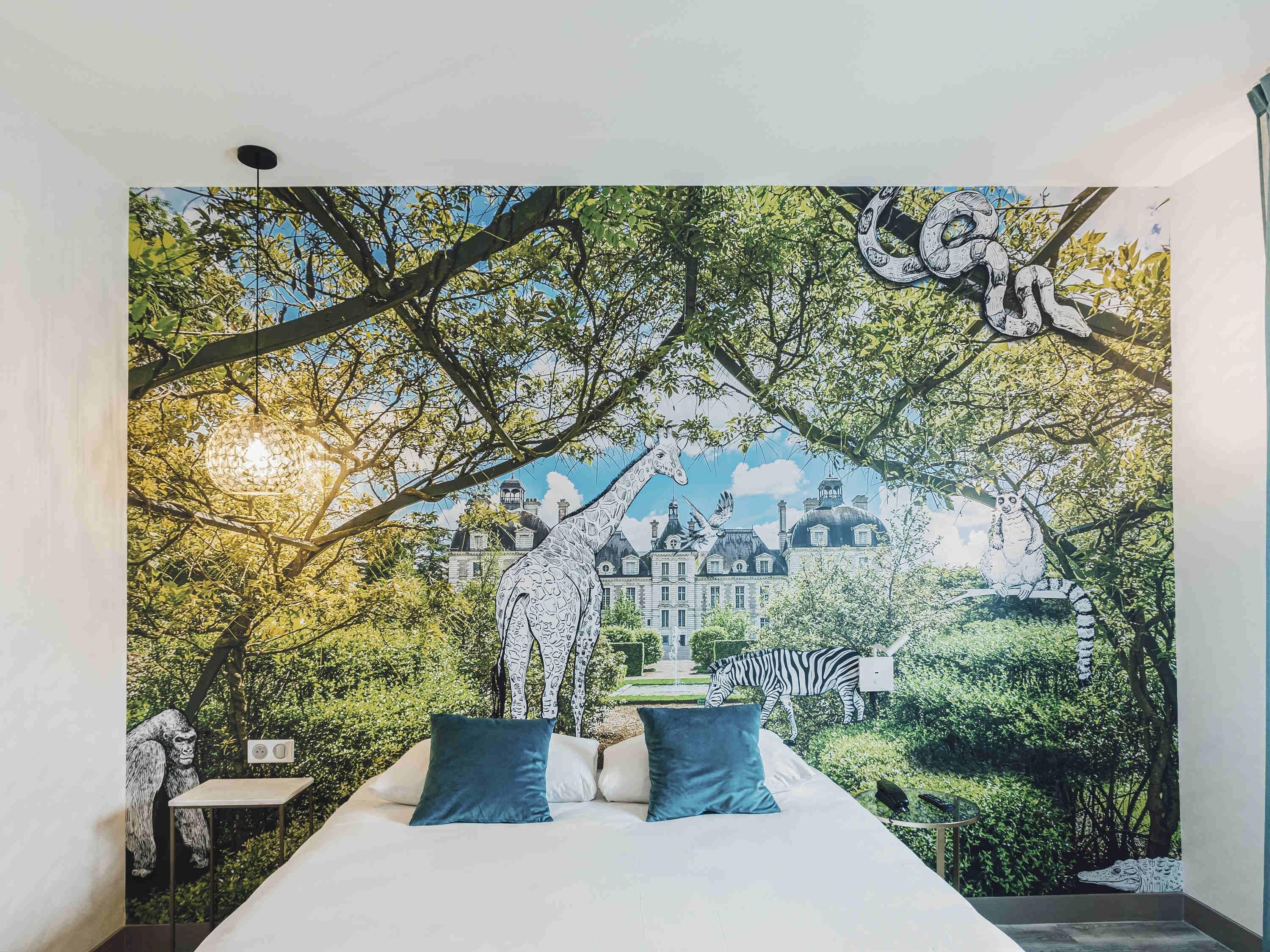 Ein Schlafzimmer mit einer Wandmalerei von einem Wald und Tieren, einem Bett mit blauen Kissen und Blick auf ein Gebäude im Hintergrund.