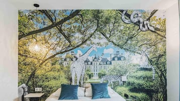 Ein Schlafzimmer mit einer Wandmalerei von einem Wald und Tieren, einem Bett mit blauen Kissen und Blick auf ein Gebäude im Hintergrund.