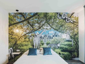 Une chambre avec une fresque représentant une forêt et des animaux, un lit avec des oreillers bleus et, au loin, une vue sur un bâtiment.