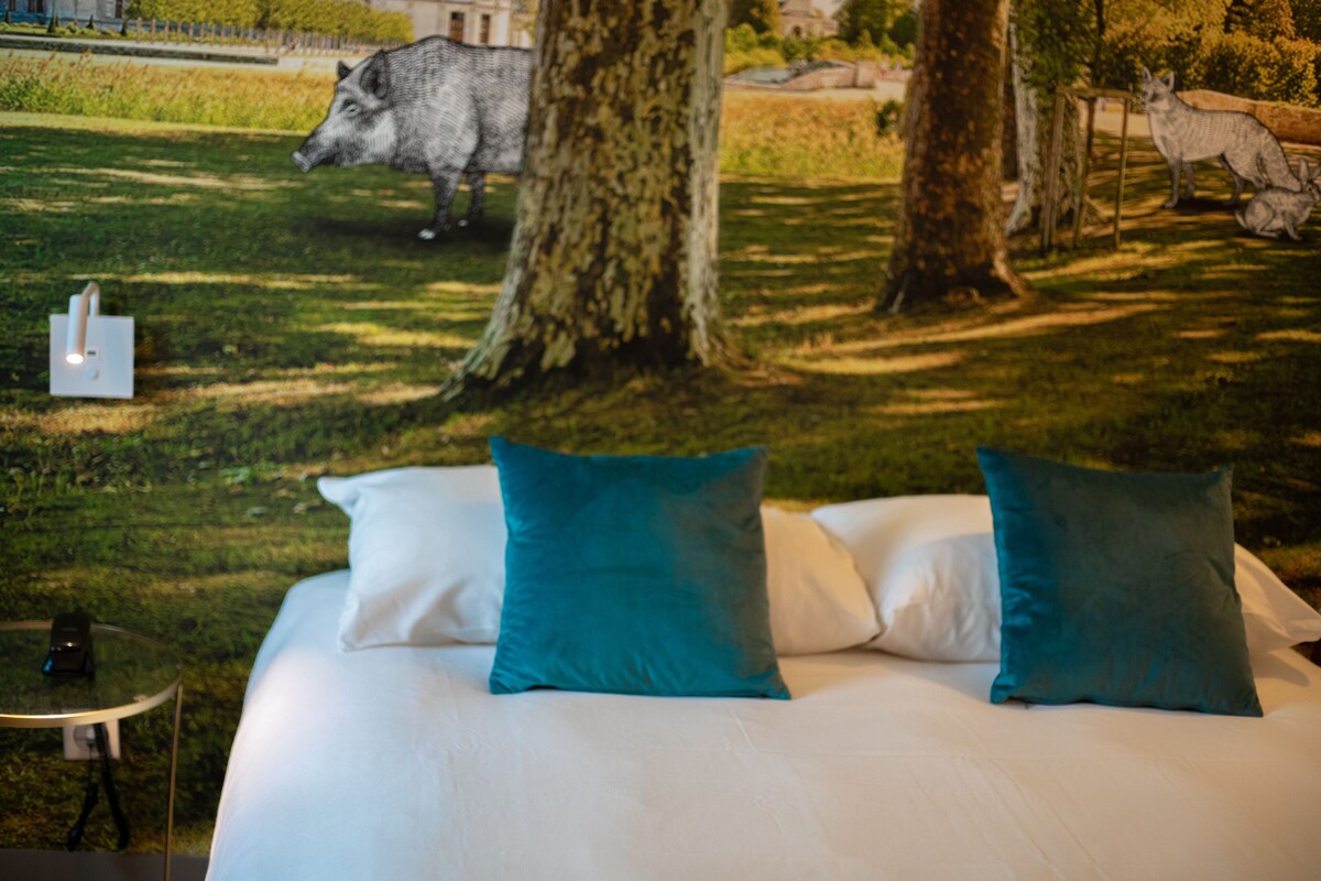 Un lit avec du linge de lit blanc et des coussins turquoise, une petite table avec un téléphone, et une sculpture de sanglier dans un espace verdoyant bordé d’arbres.