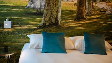 Una cama con sábanas blancas y almohadas color turquesa, una mesita con un teléfono y una escultura de un jabalí en un área con césped y árboles.