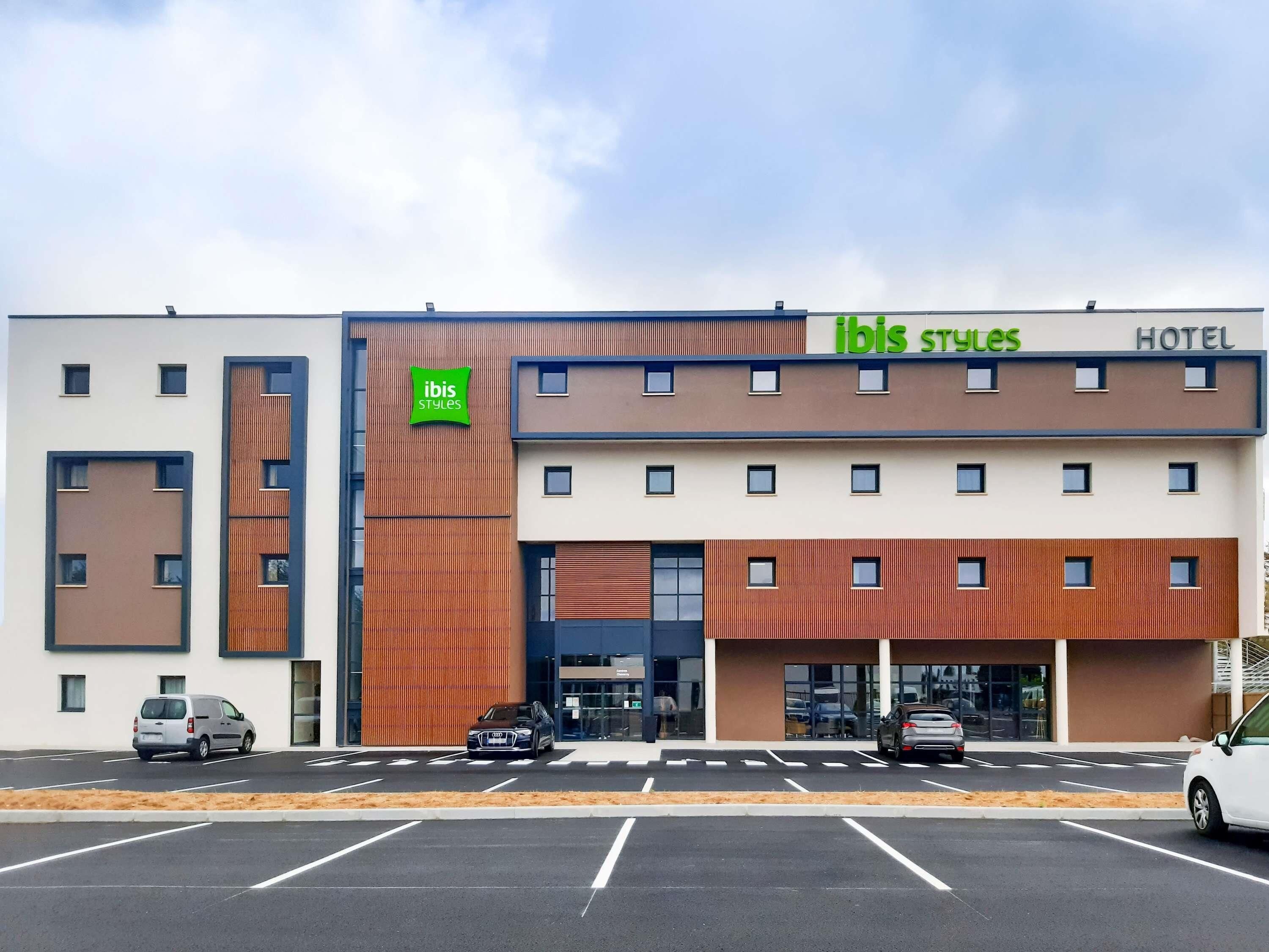 Een modern hotel met een plat dak, een opvallend groen logo en een parkeerplaats met meerdere auto's.