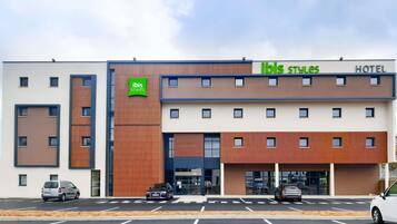 Een modern hotel met een plat dak, een opvallend groen logo en een parkeerplaats met meerdere auto's.