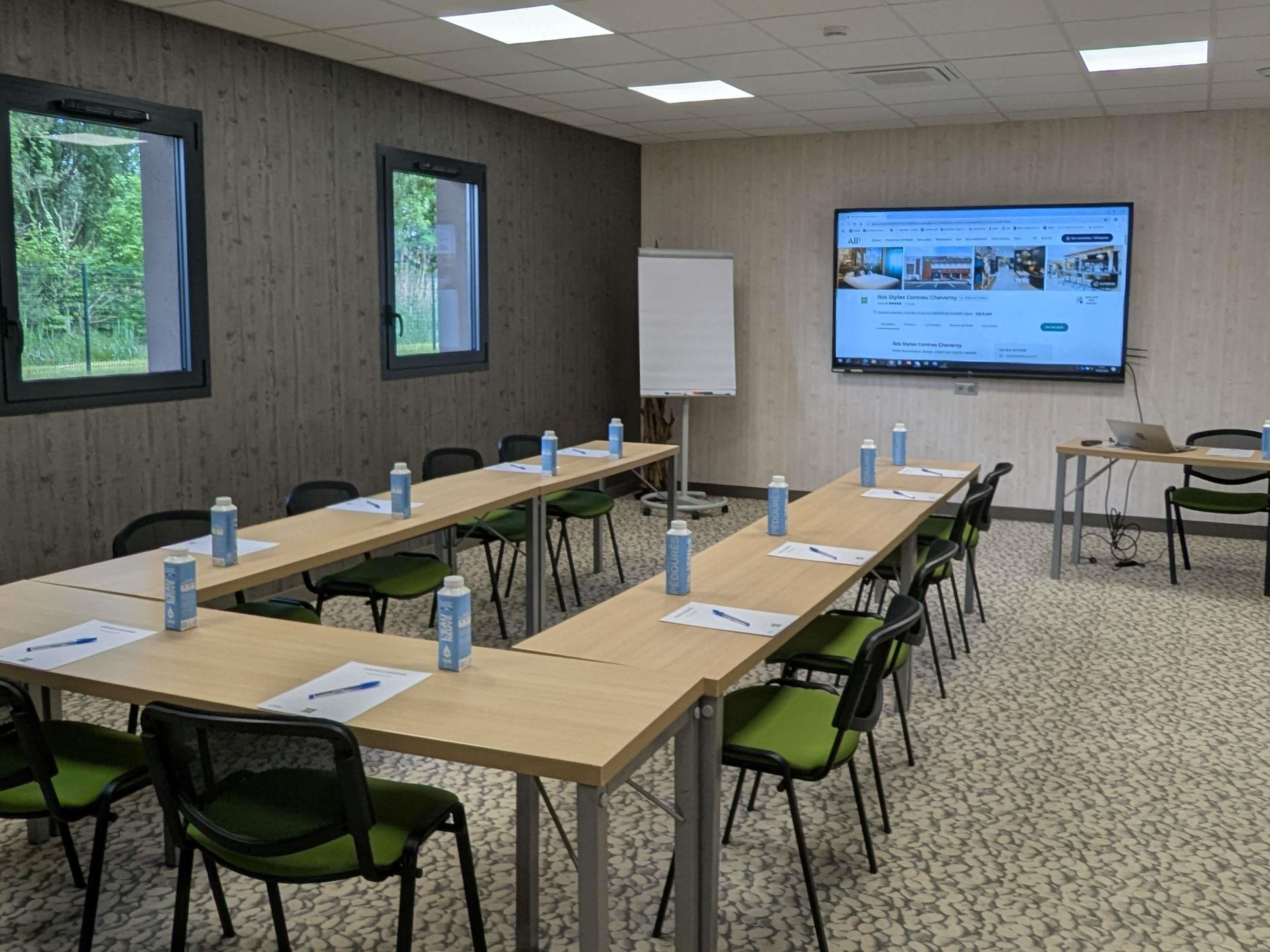 Ein Konferenzraum mit langen Tischen, grünen Stühlen, Wasserflaschen, einer Tafel und einer Projektionsleinwand, auf der eine Webseite angezeigt wird.