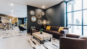 Un hall d’entrée moderne avec une grande fenêtre, un canapé en cuir marron agrémenté de coussins jaunes, une table basse en bois avec une plante, et des miroirs décoratifs fixés au mur.