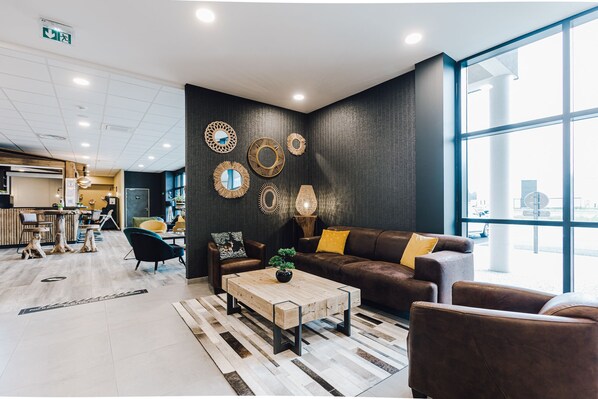 Un hall d’entrée moderne avec une grande fenêtre, un canapé en cuir marron agrémenté de coussins jaunes, une table basse en bois avec une plante, et des miroirs décoratifs fixés au mur.