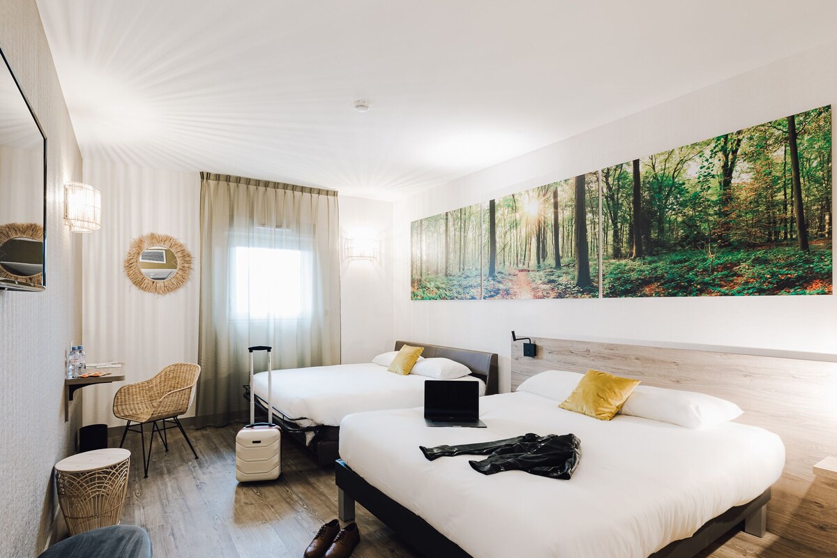 Une chambre d’hôtel avec deux lits, un bureau, une chaise, une valise et un grand tableau sur le thème de la forêt accroché au mur.
