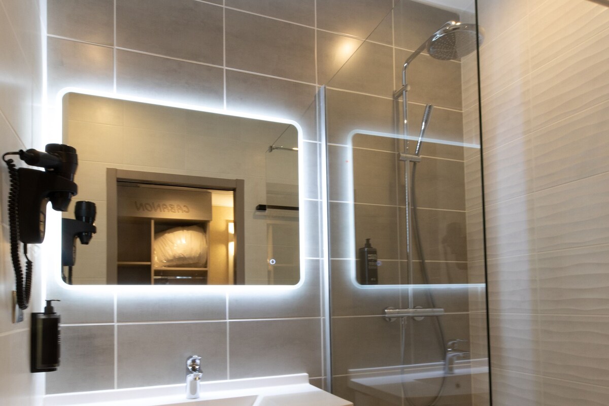 Une salle de bain moderne dotée d’une douche à parois de verre, d’un meuble-lavabo avec miroir et d’un sèche-cheveux fixé au mur.