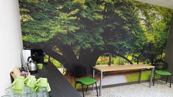 Un área de recepción con un escritorio de madera, sillas verdes, una máquina de café y un mural grande con árboles.