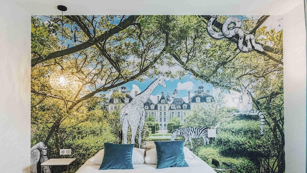 Une chambre avec une fresque représentant une forêt et des animaux, un lit avec des oreillers bleus et, au loin, une vue sur un bâtiment.