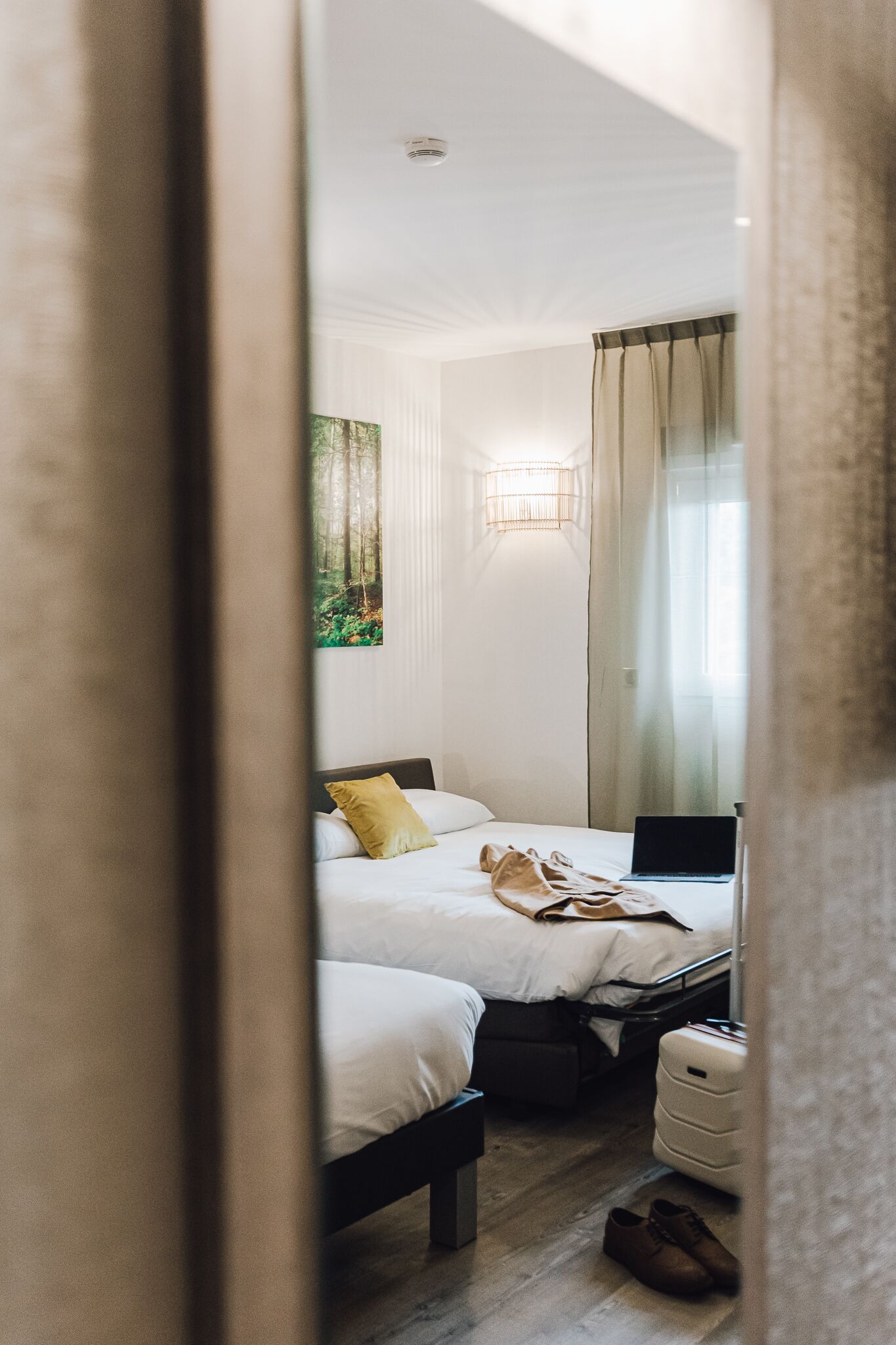 A bedroom with two beds, a bedside table, a lamp, a painting on the wall, and a window with curtains.