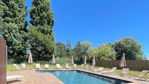 Une piscine rectangulaire, agrémentée de chaises longues et de parasols, entourée d’une terrasse en bois et de verdure.