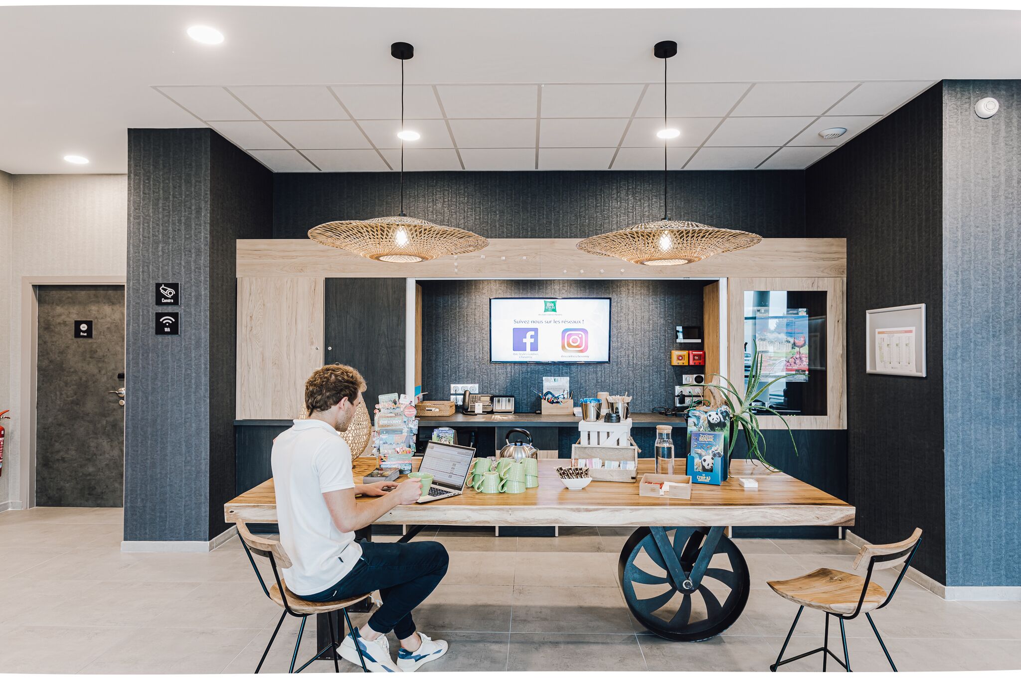 A modern reception area with a wooden reception desk, two hanging wicker lamps, a digital screen displaying social media icons, and a variety of items on the desk including a laptop, a plant, and a coffee machine.