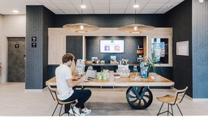 A modern reception area with a wooden reception desk, two hanging wicker lamps, a digital screen displaying social media icons, and a variety of items on the desk including a laptop, a plant, and a coffee machine. - ibis Styles Contres Cheverny (Le Controis en Sologne)