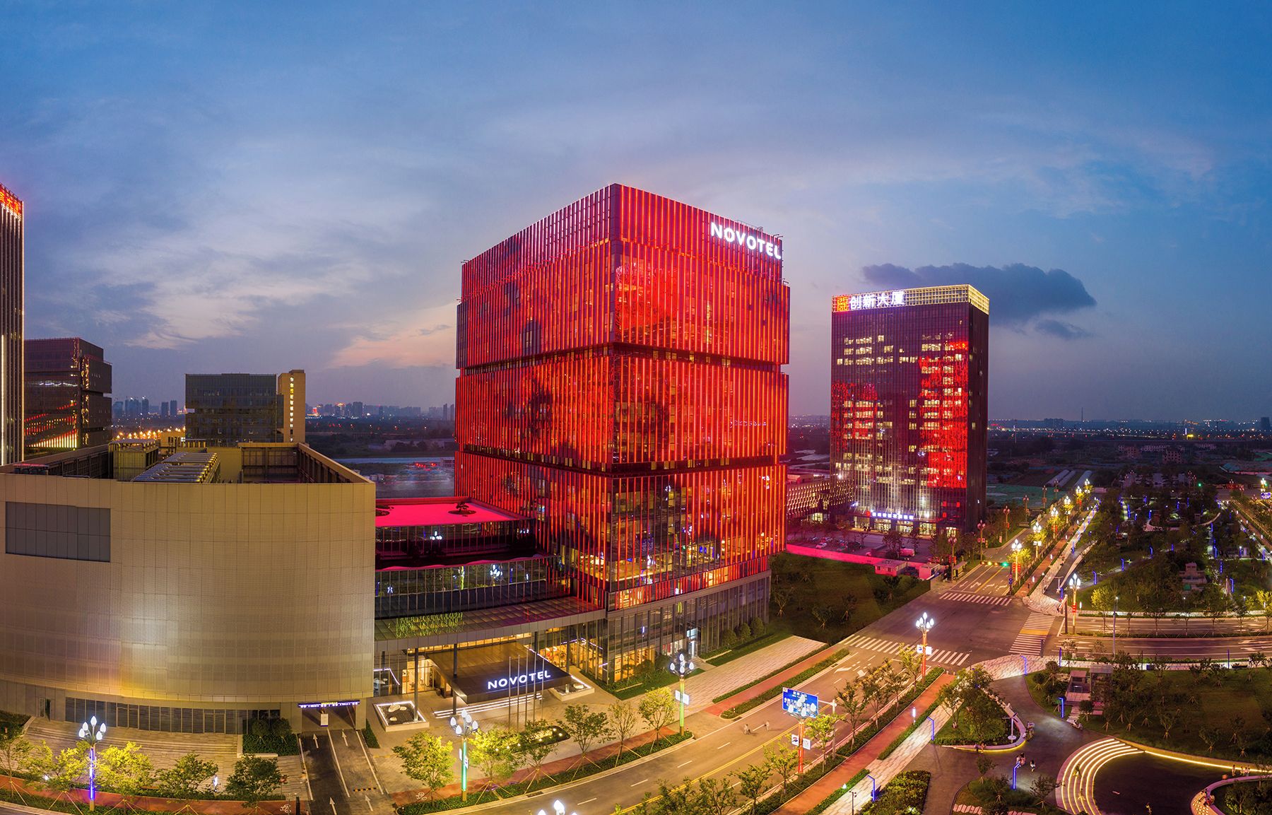 Un hotel moderno con una fachada roja destacada, una entrada bien iluminada y vistas del paisaje urbano circundante.