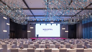 A conference room with rows of tables, a large screen displaying 'NOVOTEL,' and a modern chandelier.