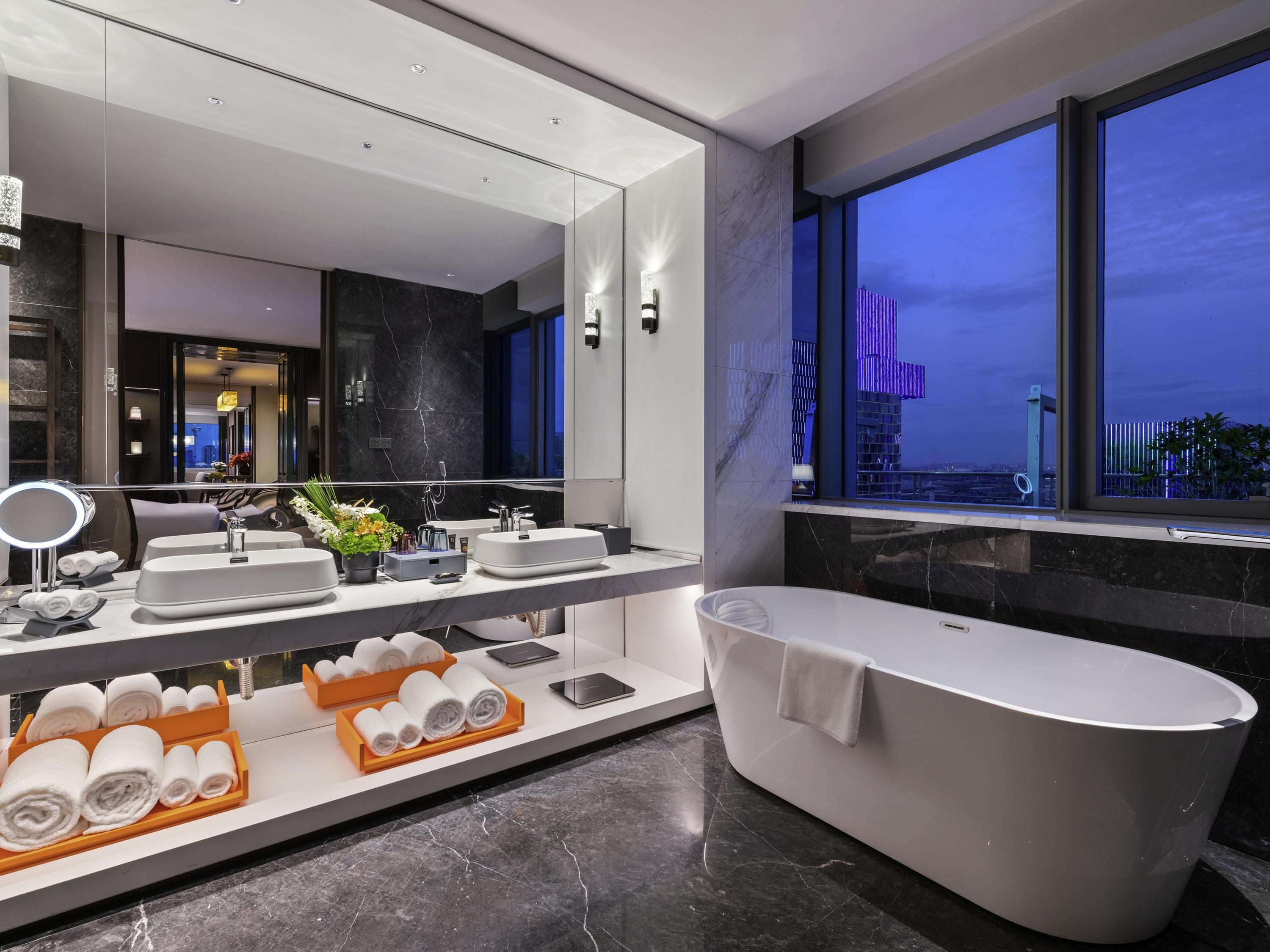 A modern bathroom with a large mirror, two sinks, a bathtub, and a view of the city at dusk.